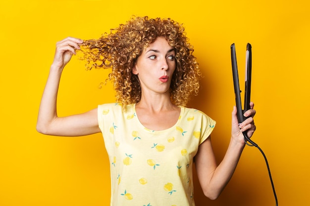 Jovem com cabelo encaracolado parece surpresa com o alisador de cabelo em um fundo amarelo