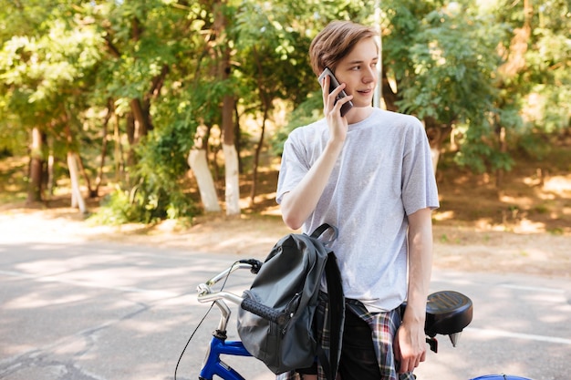 Jovem com cabelo loiro em pé com mochila e bicicleta e sonhadoramente olhando de lado enquanto fala em seu telefone Retrato de menino sorridente em pé e usando seu celular enquanto passa o tempo no parque