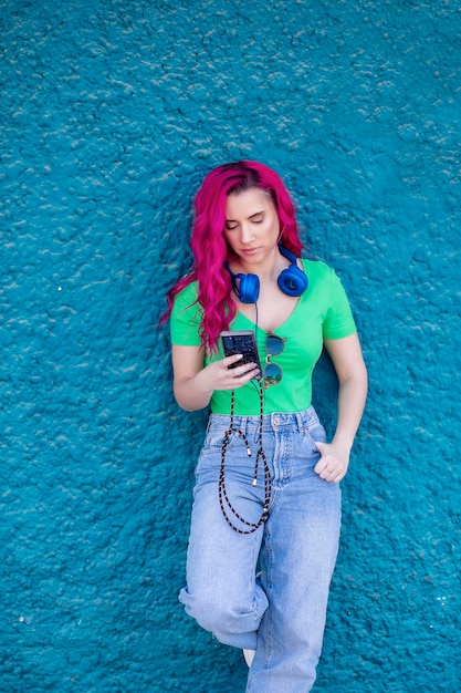 Jovem com cabelo rosa encostado em uma parede colorida e ouvindo música com seus fones de ouvido