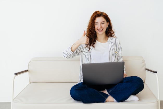 Jovem com cabelo ruivo encaracolado está deitada no sofá e participando de uma videoconferência em um laptop freelance e trabalho remoto