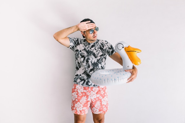 Jovem com camisa floral, boné e óculos no verão com um patinho flutuando fazendo gestos de calor, à luz do dia em uma parede branca