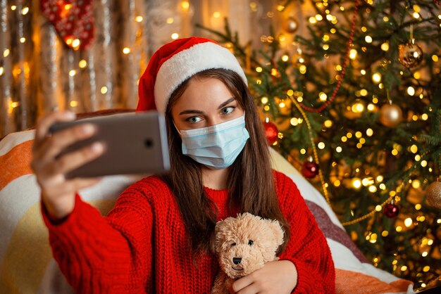 Jovem com chapéu de natal sentada na poltrona e tirando selfie pelo smartphone