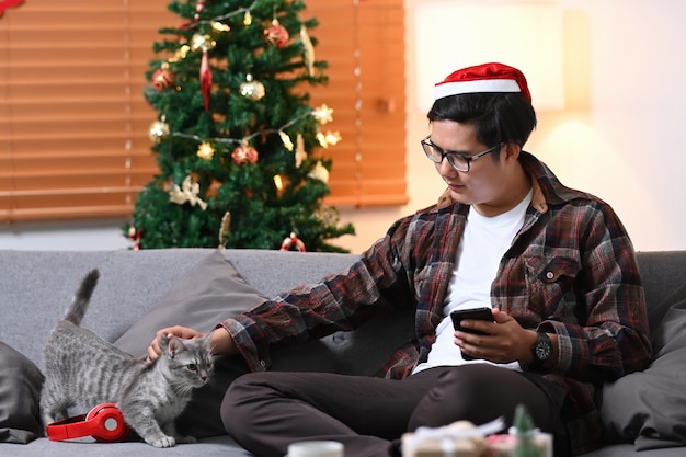 Jovem com chapéu de Papai Noel sentado em um sofá com seu gato em casa.