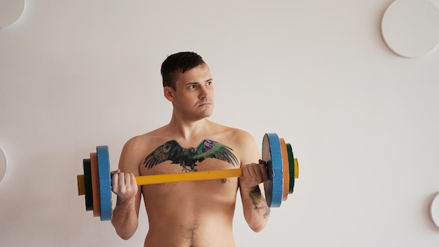 Jovem com corpo nu e tatuagem no peito levantando barra multicolorida contra parede branca estampada Cara forte adulto fazendo esporte em casa
