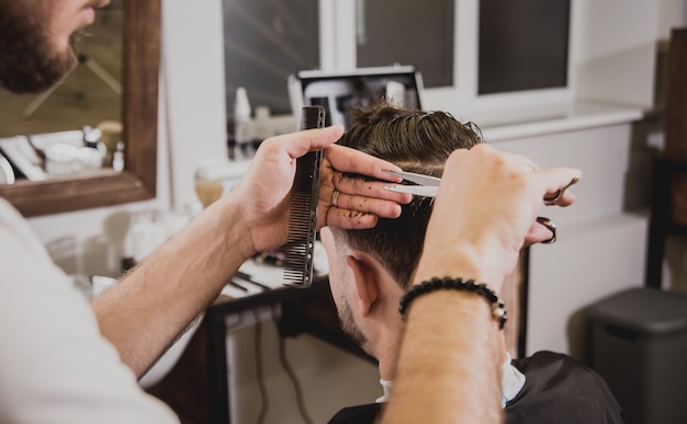 Jovem com corte de cabelo na moda na barbearia. O barbeiro faz o penteado e a barba.