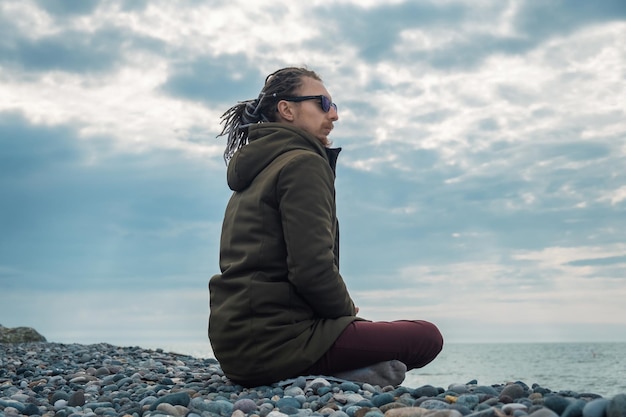 Jovem com dreadlocks praticando ioga meditação está sentado em posição de lótus ao ar livre à beira-mar Saúde mental