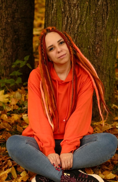 Jovem com dreadlocks sentado perto da árvore no outono dourado Mulher vívida com penteado colorido posando na natureza