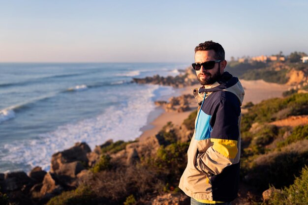 Jovem com jaqueta e óculos de sol olhando para o mar de um penhasco ao lado da praia lazer e relaxar espaço de cópia de conceito para texto