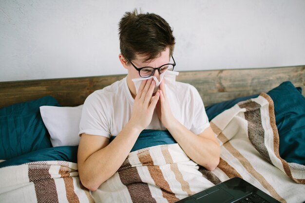 Jovem com lenço. o cara doente na cama tem nariz escorrendo. homem faz uma cura para o resfriado comum