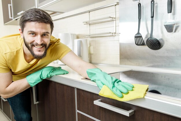 Jovem com luvas protetoras verdes limpando a cozinha moderna com pano amarelo