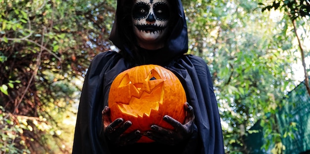 Foto jovem com máscara facial de pintura de halloween usando capuz preto bruxa assustadora segurando abóbora cravada assustadora