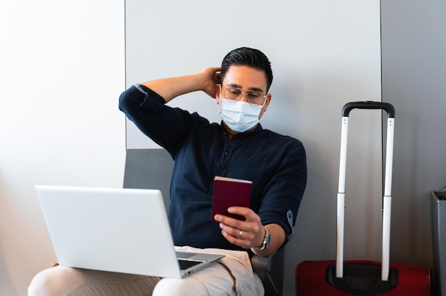 Jovem com máscara facial sentado na sala de espera no aeroporto Empresário usando computador