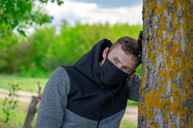 Jovem com máscara médica preta e luvas encostado na árvore na floresta Macho adulto desfrutando da natureza no campo no período de infecção epidêmica por coronavírus