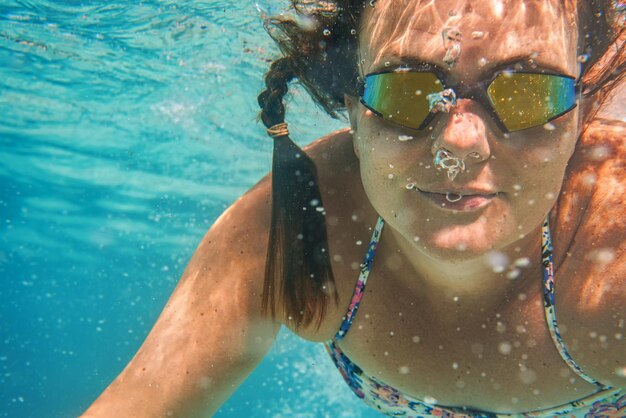 Foto jovem com óculos de natação reflexivos nada sob a água no mar, bolhas iluminadas pelo sol sobre o rosto, detalhes subaquáticos