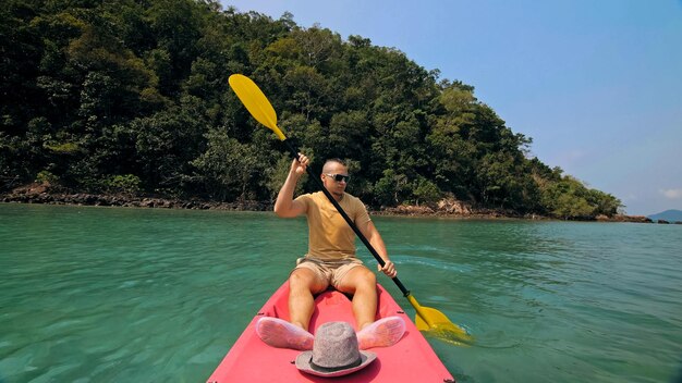 Jovem com óculos de sol e chapéu enfileira canoa de plástico rosa ao longo do mar contra ilhas montanhosas verdes com selvas selvagens Viajando para países tropicais Cara forte está navegando em caiaque em vista frontal para o mar