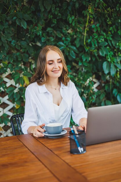 Jovem com roupas casuais, tomando café e falando ao telefone, trabalhando em um terraço verde em um laptop, olhando para a tela e trabalhando em um projeto remotamente do trabalho em um café