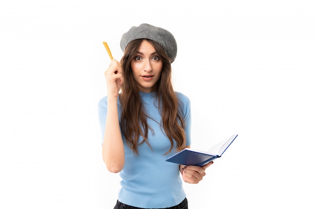 Jovem com sorriso encantador, cabelo castanho longo ondulado, maquiagem linda, em jersey azul, jeans preto, boina cinza, em pé com caneta e caderno