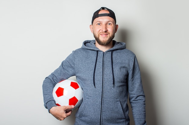 Jovem com um boné e um moletom segura uma bola de futebol contra uma parede de luz.