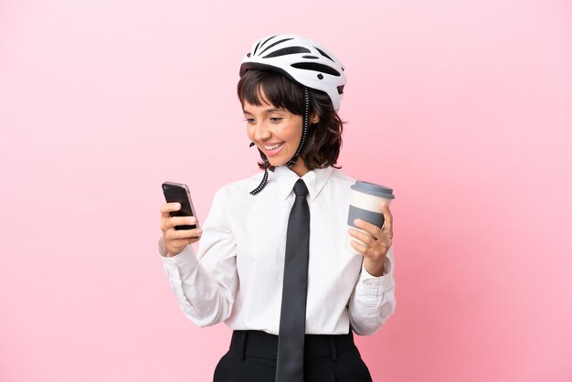 Jovem com um capacete de bicicleta isolado em fundo rosa segurando café para levar e um celular