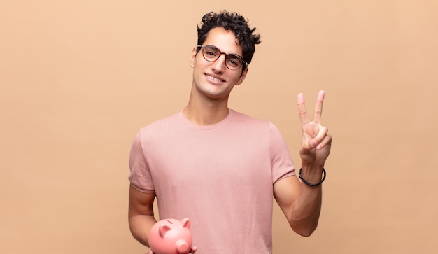 Jovem com um cofrinho sorrindo e parecendo feliz, despreocupado e positivo, gesticulando vitória ou paz com uma mão