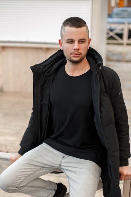 Jovem com um penteado curto em uma jaqueta preta de inverno e jeans branco sentado na rua