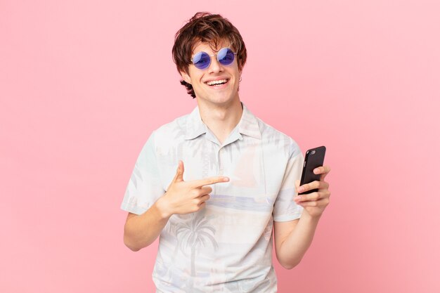 Jovem com um telefone celular sorrindo alegremente se sentindo feliz e apontando para o lado