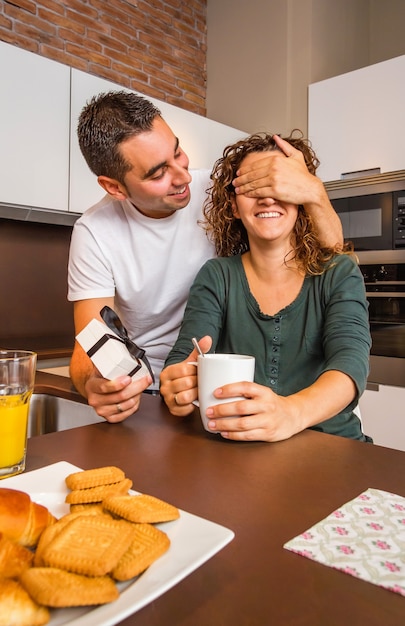 Jovem com uma caixa de presente fechando os olhos da namorada para uma surpresa enquanto toma café da manhã na cozinha da casa