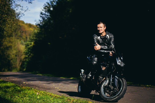 jovem com uma jaqueta de couro preta sentado em uma motocicleta esportiva no fundo de uma bela estrada na floresta