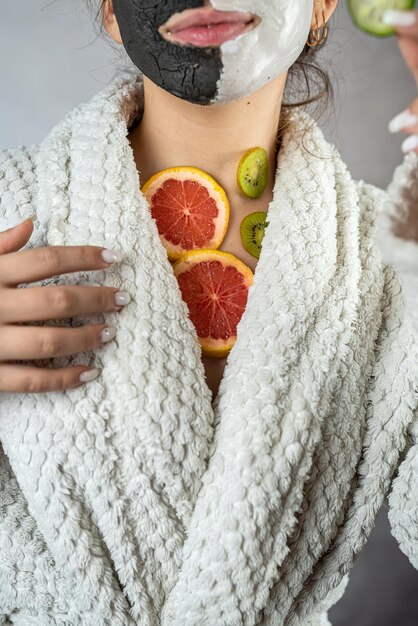 Jovem com uma máscara de argila preta e branca no rosto está segurando frutas frescas de toranja kiwi