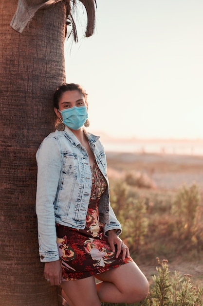 Jovem com uma máscara facial na praia ao pôr do sol sob um retrato de palmeira