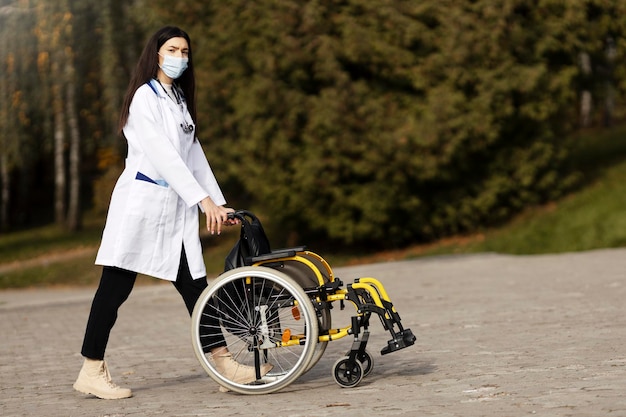 Jovem com uma máscara protetora e bata médica carrega uma cadeira de rodas Enfermeira atraente olha para a câmera carrega uma cadeira de rodas ao ar livre