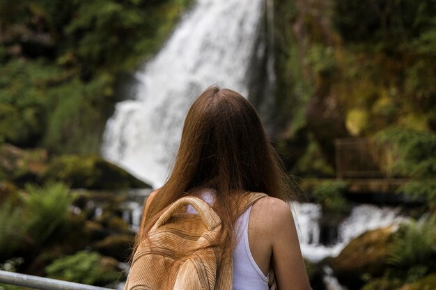 Foto jovem com uma mochila parada de costas olhando para uma cachoeira