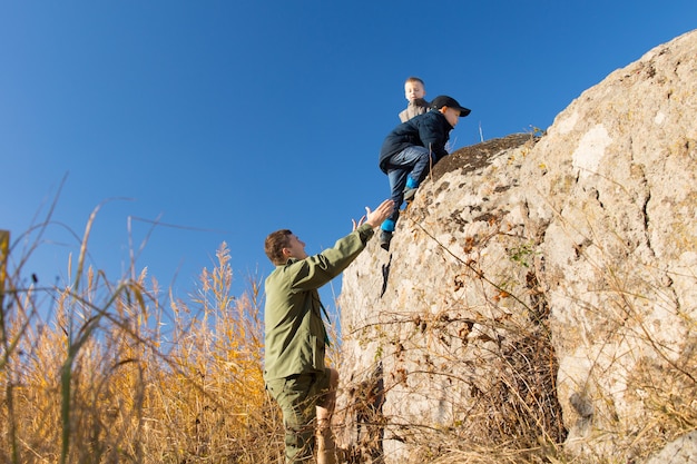 Jovem com uniforme de escuteiro ajudando dois meninos a escalar um penhasco rochoso no deserto, apoiando-os com a mão lá embaixo