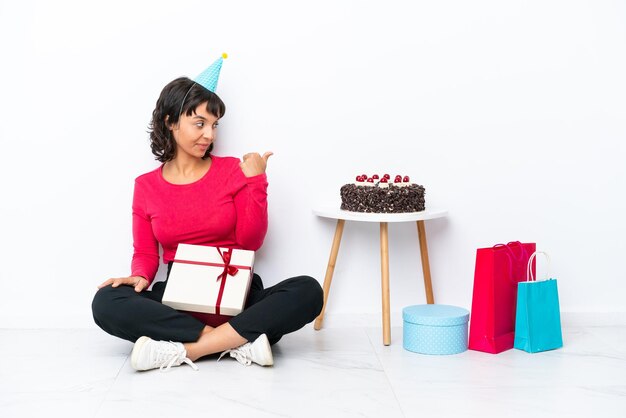 Jovem comemorando seu aniversário sentado no chão isolado no fundo branco apontando para o lado para apresentar um produto