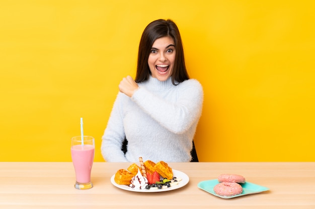 Jovem comendo waffles e milk-shake em uma mesa ao longo da parede amarela isolada comemorando uma vitória