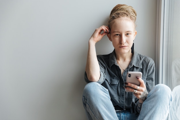 Foto jovem concentrada com dreadlocks e piercing dentro de casa usando telefone celular perto da janela.