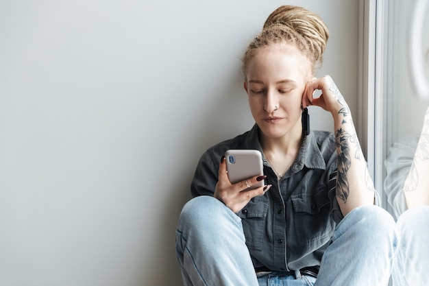 jovem concentrada com dreadlocks e piercing dentro de casa usando telefone celular perto da janela.