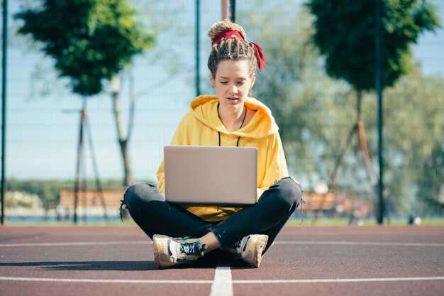 Jovem concentrada olhando pensativamente para a tela de seu laptop enquanto está sentada no meio de um campo de esportes