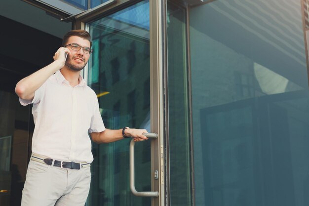 Jovem confiante abrindo a porta e saindo do prédio de escritórios moderno enquanto fala no celular com colegas sobre reunião, copie o espaço. Conceito de negócios, tecnologia e comunicação