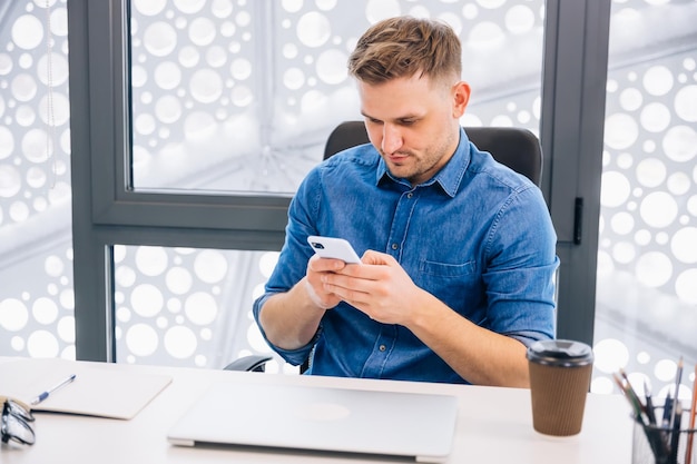 Jovem confiante em roupas casuais elegantes segurando smartphone em seu local de trabalho no escritório