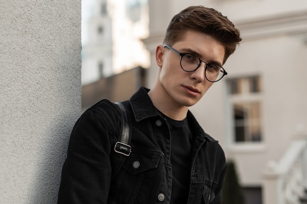 Jovem confiante urbano retrato fresco com penteado na jaqueta jeans preta da moda em óculos elegantes perto da parede ao ar livre. Modelo de cara bonito na moda descansando na cidade. Moda masculina casual. Estilo de rua.