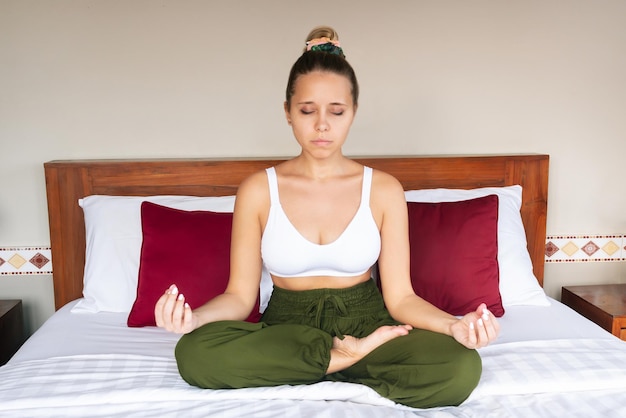 Jovem consciente medita segurando os dedos em sinal de ioga com os olhos fechados sentado na cama