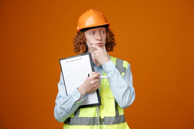 Jovem construtor em uniforme de construção e capacete de segurança segurando a prancheta e marcador olhando de lado com expressão pensativa pensando em pé sobre fundo laranja