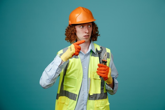 Jovem construtor em uniforme de construção e capacete de segurança segurando chave e alicate olhando de lado preocupado apontando com o dedo indicador para o lado em pé sobre fundo azul