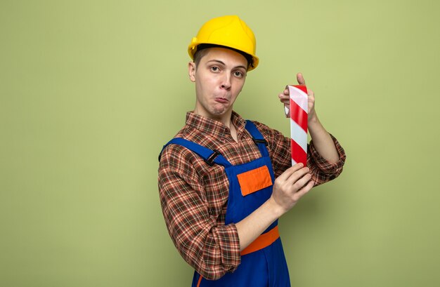 Jovem construtor impressionado usando uniforme segurando fita adesiva