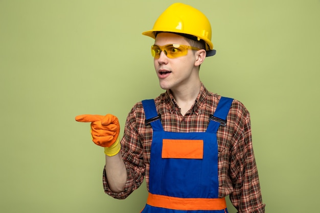 Jovem construtor vestindo uniforme e luvas com óculos, isolado em parede verde oliva com espaço de cópia