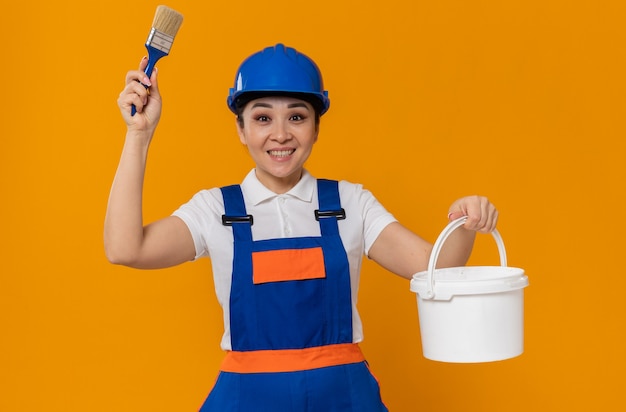 Jovem construtora asiática impressionada com capacete de segurança azul segurando pincel e tinta a óleo