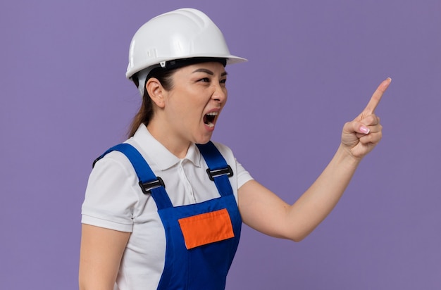 Jovem construtora asiática irritada com capacete de segurança branco olhando e apontando para o lado gritando com alguém