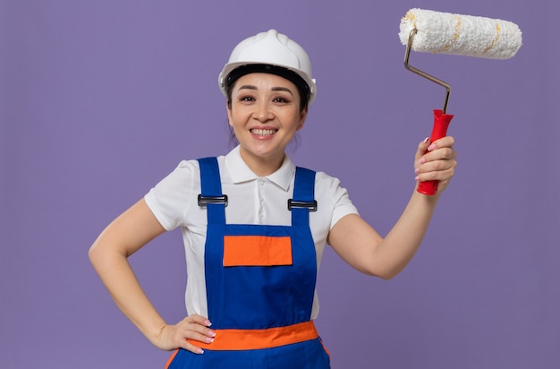 Jovem construtora asiática sorridente com capacete de segurança branco, colocando a mão em sua cintura e segurando o rolo de pintura
