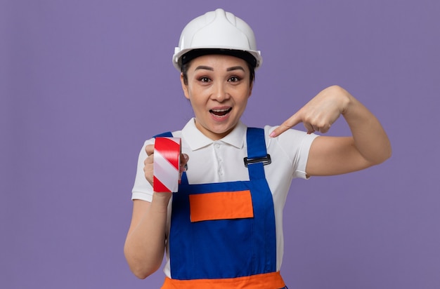 Jovem construtora asiática surpresa com capacete de segurança branco segurando e apontando para a fita de advertência